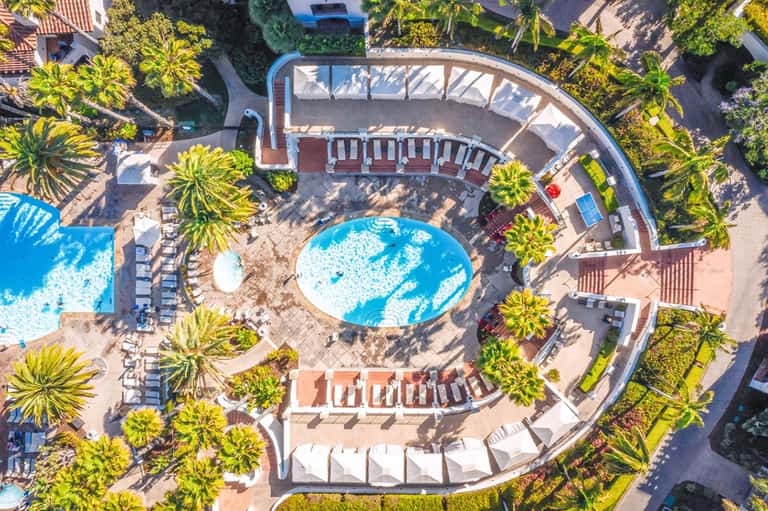 Aerial view of resort with curved pool, palm trees, lounge chairs, and surrounding buildings