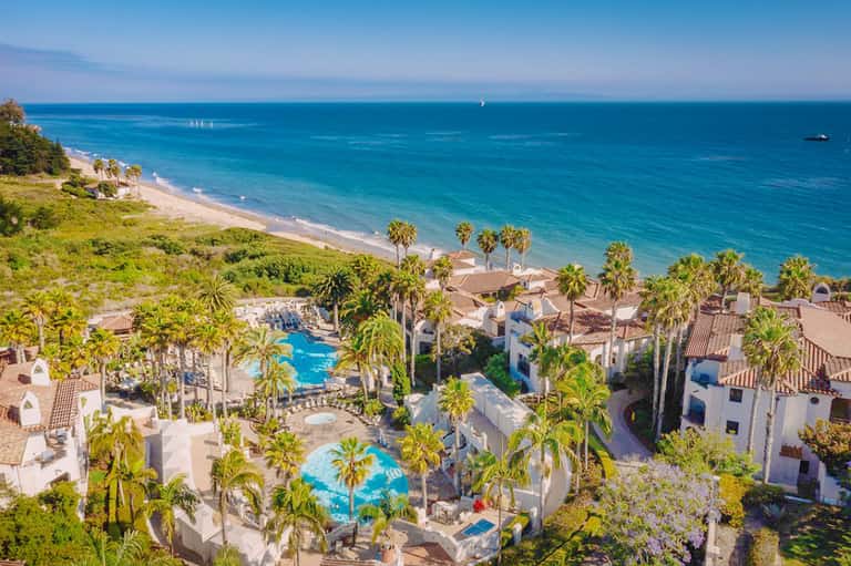 Aerial view of beachfront resort with pools, white buildings, palm trees, and Mediterranean sea
