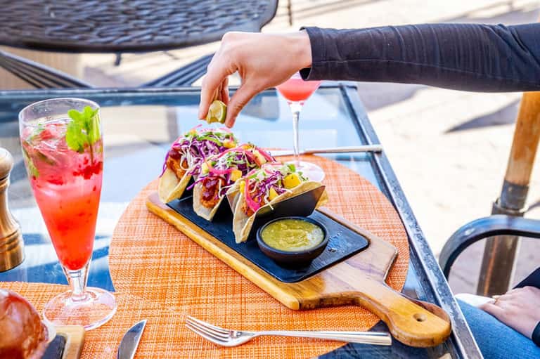 Beachside dining table with tacos, cocktails, and person reaching for lime garnish