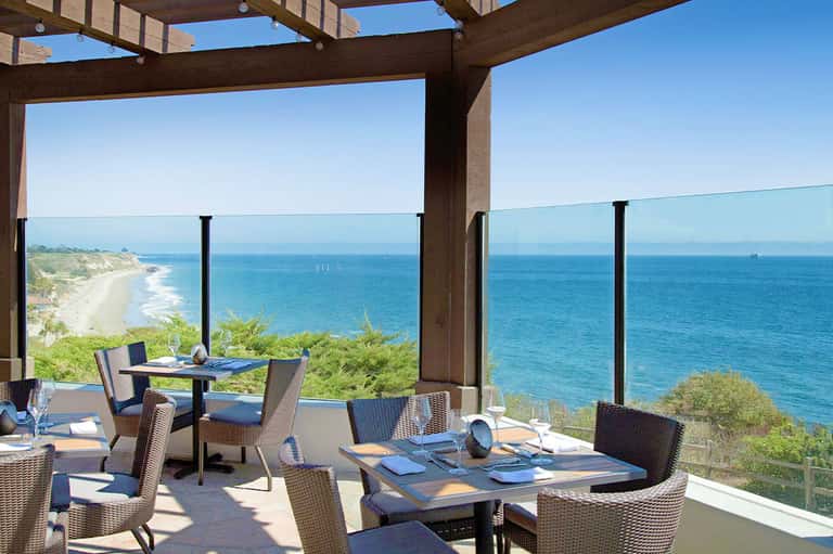 Coastal restaurant terrace with ocean and beach views, wooden pergola, modern furniture overlooking cliffs
