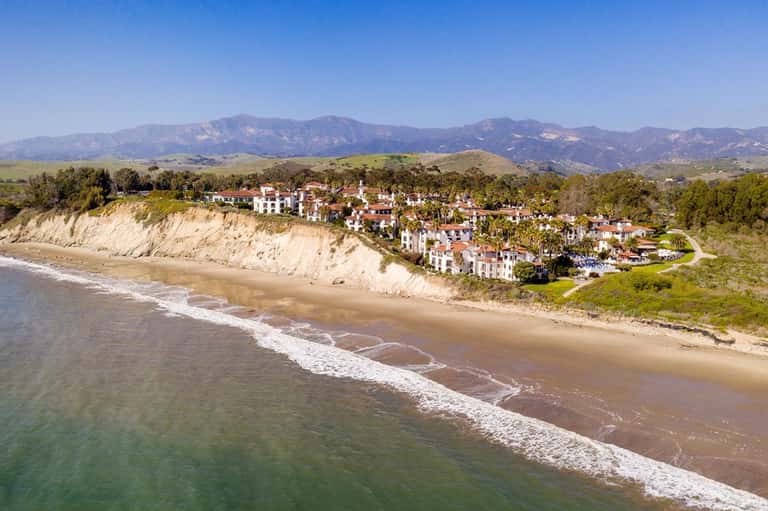 Coastal hillside resort with white buildings overlooking sandy beach and rolling mountains