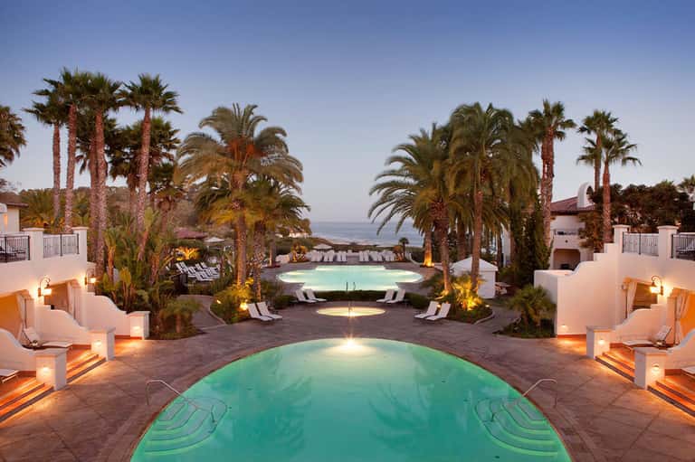 Illuminated resort pools at dusk surrounded by palm trees, loungers, and white buildings