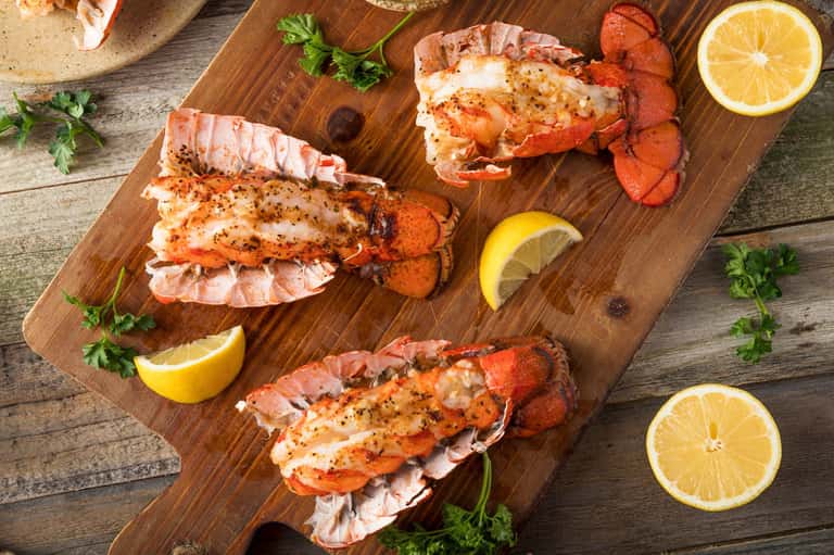 Three prepared lobster tails on wooden board with lemon slices and fresh parsley garnish