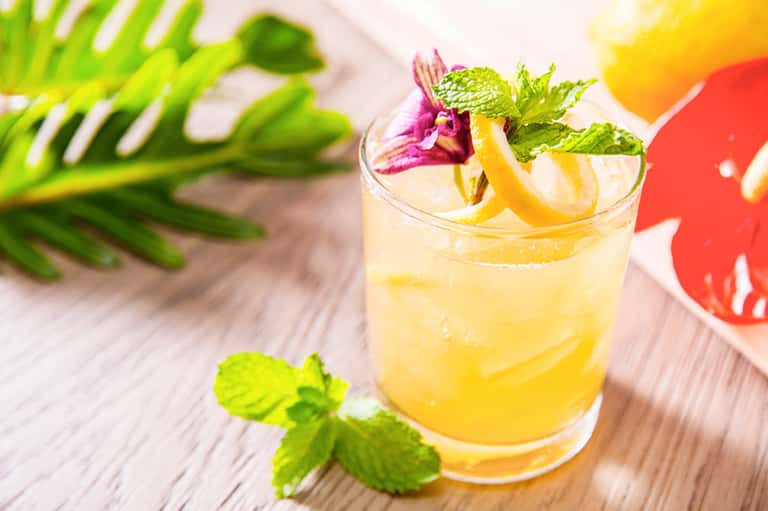 Yellow cocktail with lemon twist, mint leaves, and purple flower garnish on wooden surface