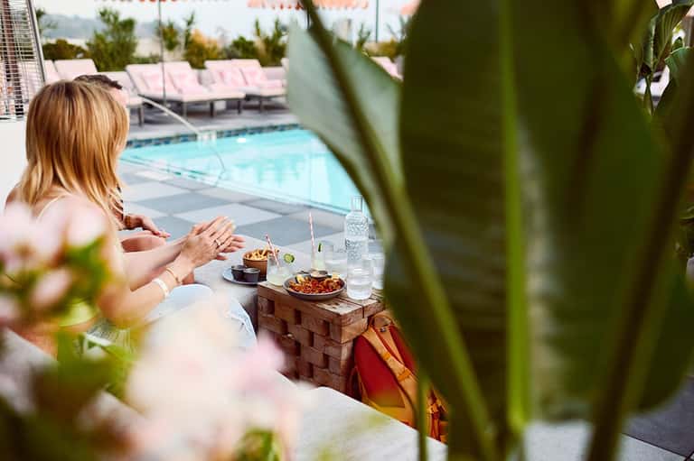 Woman relaxing poolside with food and water bottle, overlooking resort pool area with lounge chairs and green landscaping