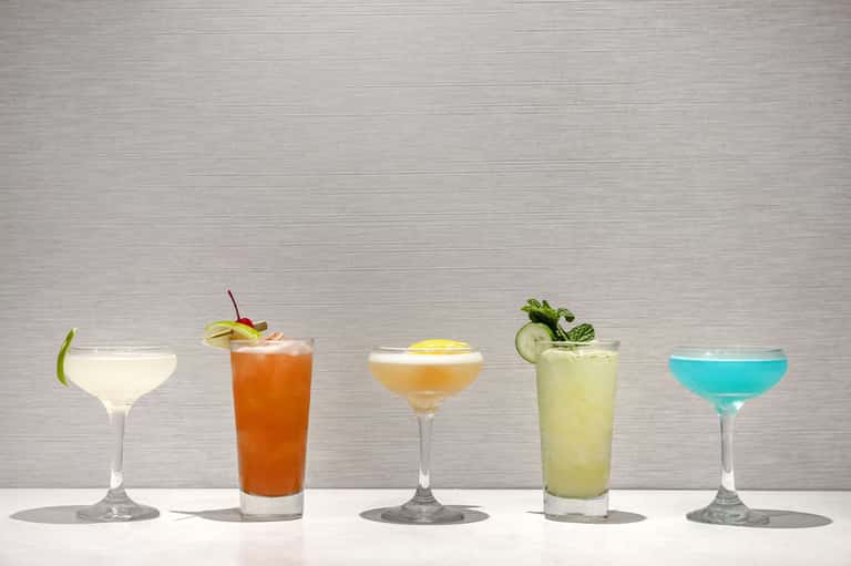 Five tropical cocktails in various colors displayed on white surface with garnishes