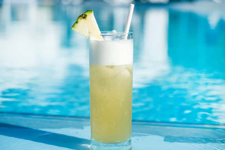 Tropical cocktail with pineapple garnish on pool deck with blurred blue water background