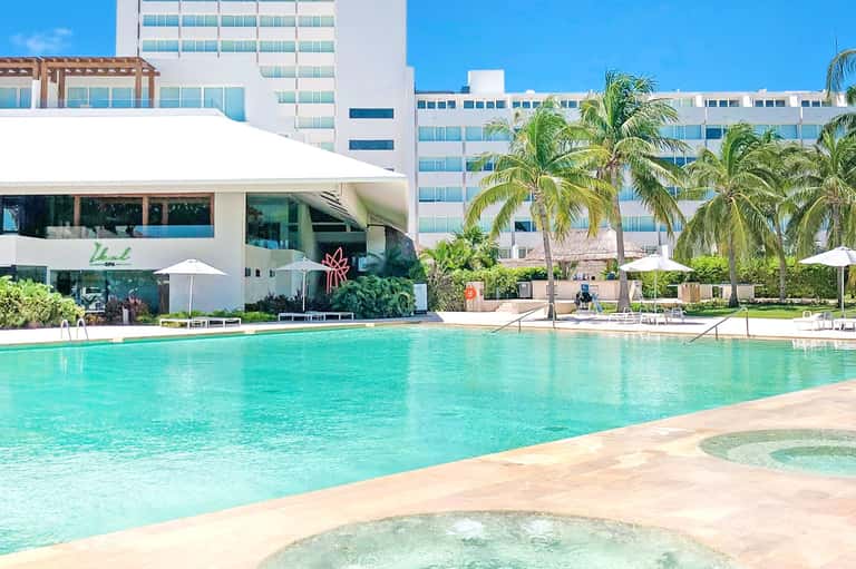 Modern resort pool area with white cabana, turquoise water, white umbrellas, and multi-story hotel buildings in background