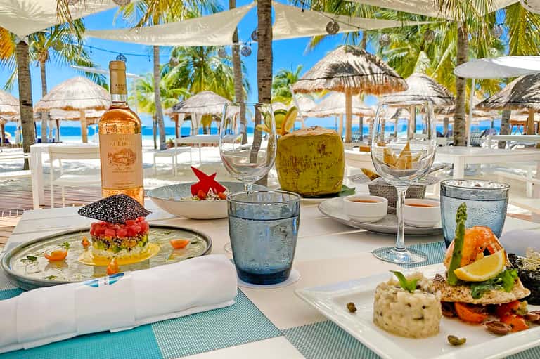 Beachfront dining table with gourmet dishes, wine, coconut drink, and ocean view with umbrellas