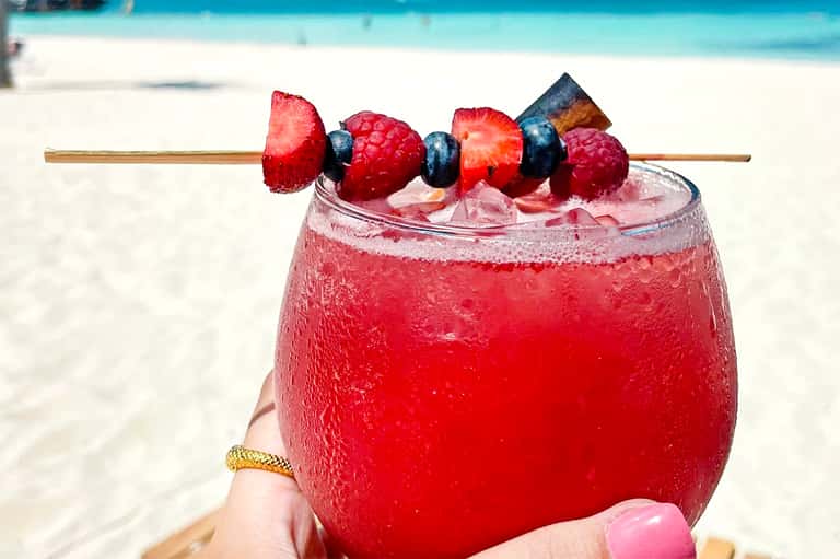Tropical cocktail with berries on skewer held against sandy beach and turquoise water