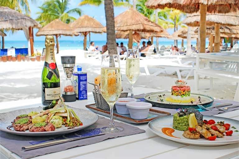 Beachfront dining table with champagne, gourmet plates, and thatched umbrellas