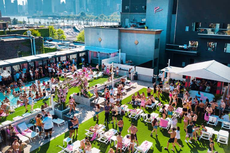 Aerial view of rooftop pool party with guests on bright green lawn and turquoise pool overlooking city skyline