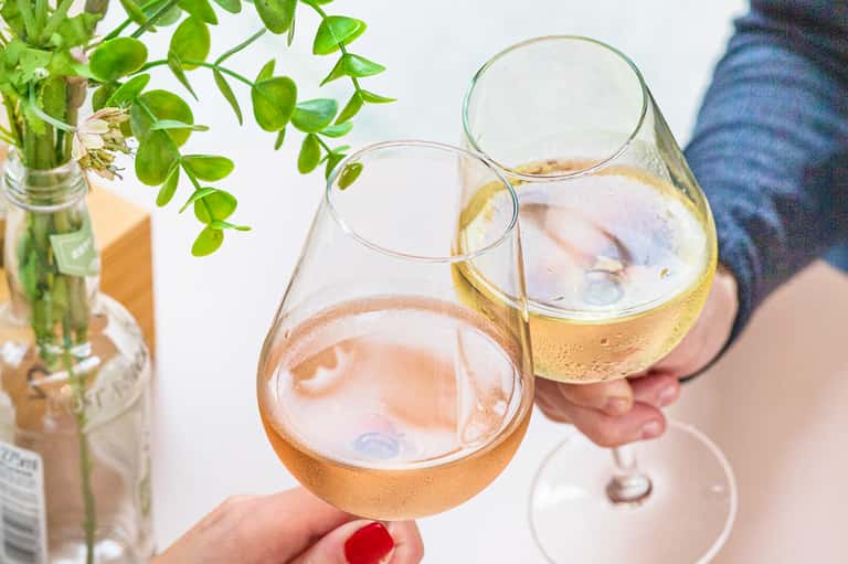 Two hands holding wine glasses with rosé and white wine outdoors near green foliage