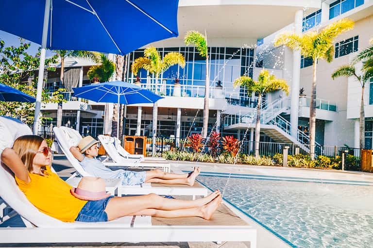Two guests lounging by pool in blue umbrellas, modern white building and tropical plants