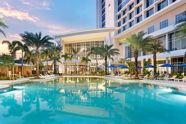 Resort pool at dusk with turquoise water, palm trees, blue umbrellas, and modern building with lit windows
