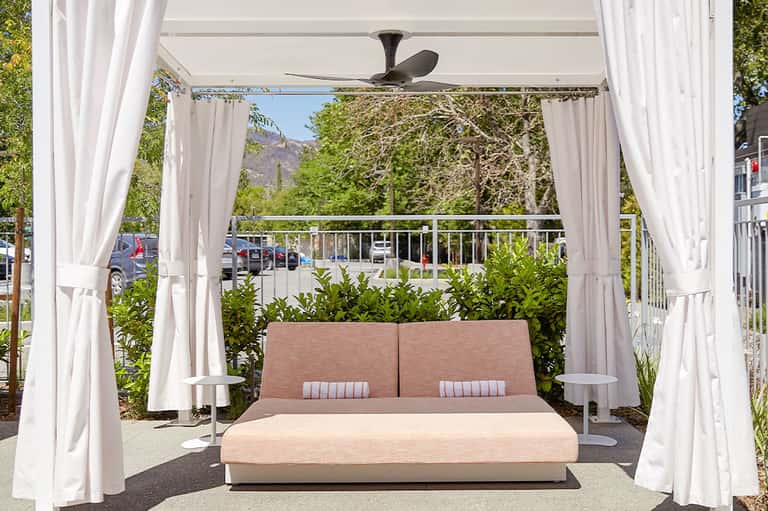 Modern outdoor lounge pavilion with white curtains, ceiling fan, pink sectional sofa, and mountain views