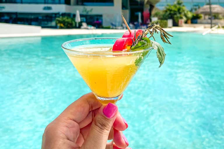 Yellow cocktail with red cherry garnish held over resort pool with cabanas in background