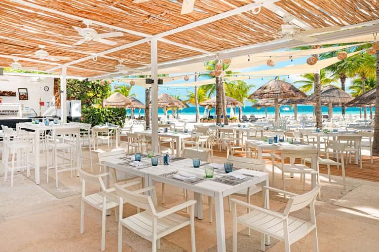 Beachfront restaurant with white furniture, wooden lattice ceiling, thatched umbrellas, and ocean beyond