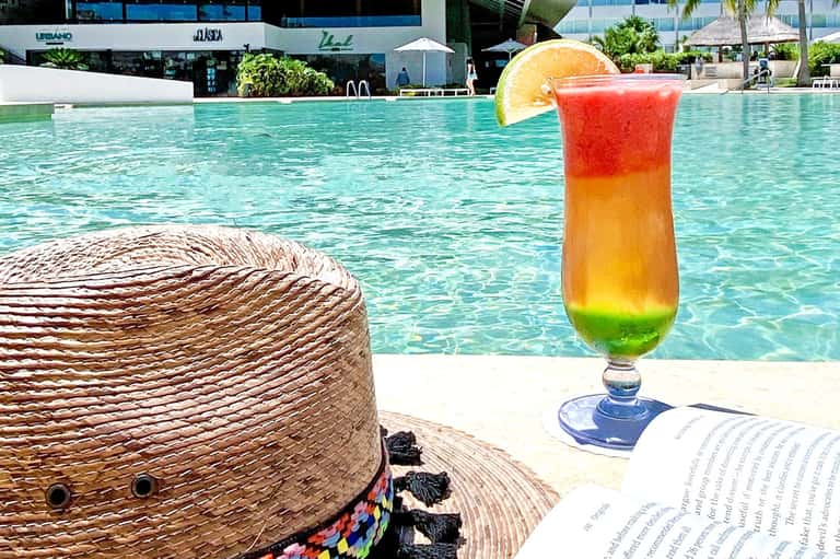 Colorful tropical drink with layered colors and orange slice near pool with straw hat