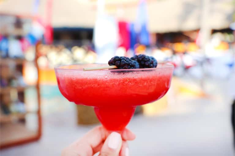 Colorful red cocktail garnished with blackberries held up against a blurred resort background