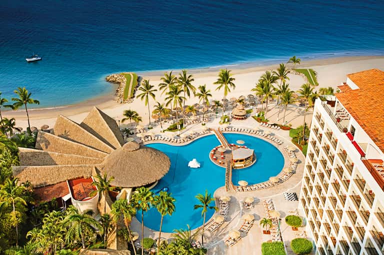 Aerial resort view with curved blue pool, thatched palapas, palm trees, pristine beach, and ocean beyond