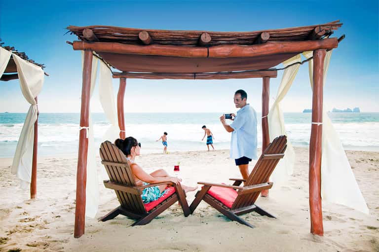 Couple relaxing in wooden beach cabana with white curtains overlooking turquoise ocean