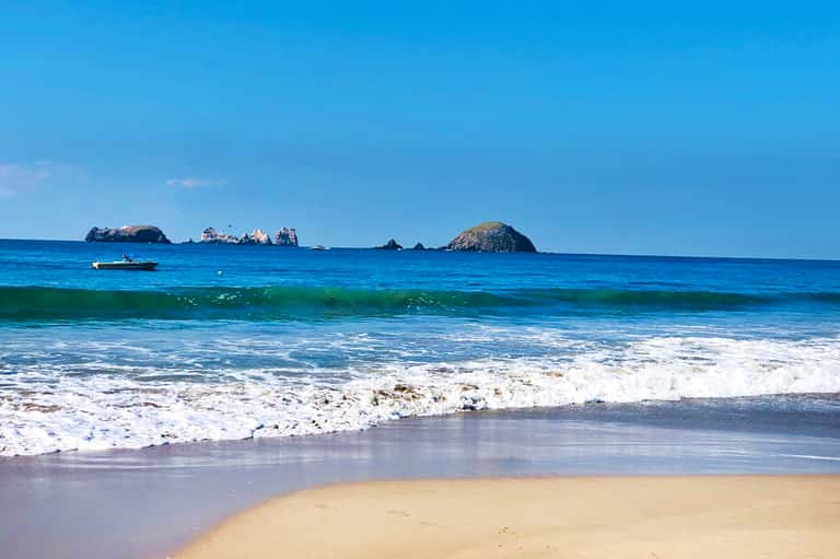 Scenic beach view with rocky islands, blue ocean, white sandy shore, and crashing waves