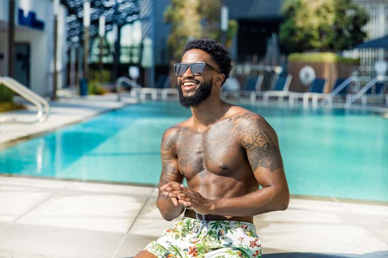 Man in sunglasses and tropical shorts smiling by resort swimming pool on sunny day
