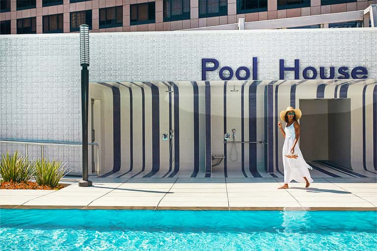 Woman in white dress and sun hat standing by pool house entrance with striped privacy screens