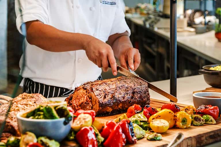 Chef carving grilled meat at resort restaurant with fresh vegetables displayed