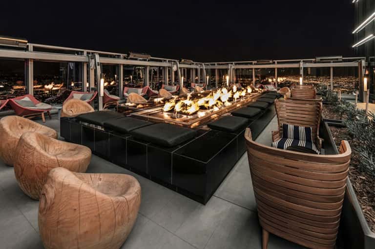 Upscale rooftop lounge at night with fire pit, wooden seating, and city lights view