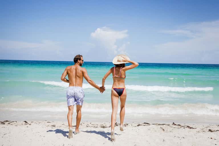 Couple holding hands running into turquoise ocean waves on sandy beach