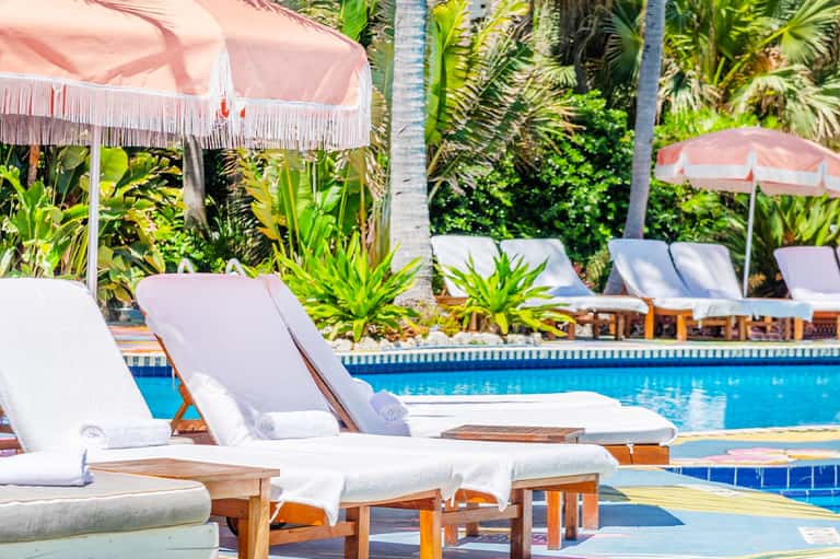 Poolside lounge chairs with pink umbrellas surrounded by tropical palm trees and vegetation