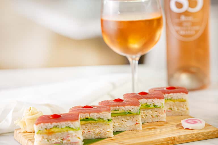 Gourmet tea sandwiches on wooden board with rosé wine and bottle on white surface