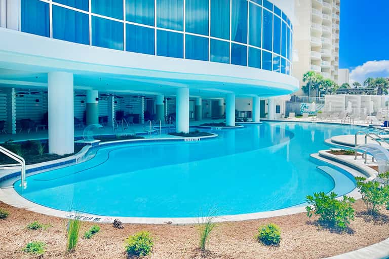 Curved resort pool with turquoise water, glass building, white columns, and ocean view under blue sky