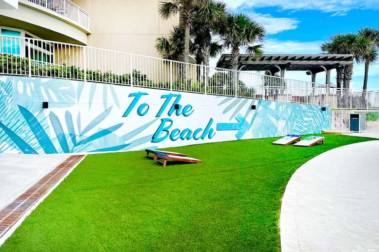 Lawn game area with cornhole boards and beach-themed mural directing to the beach