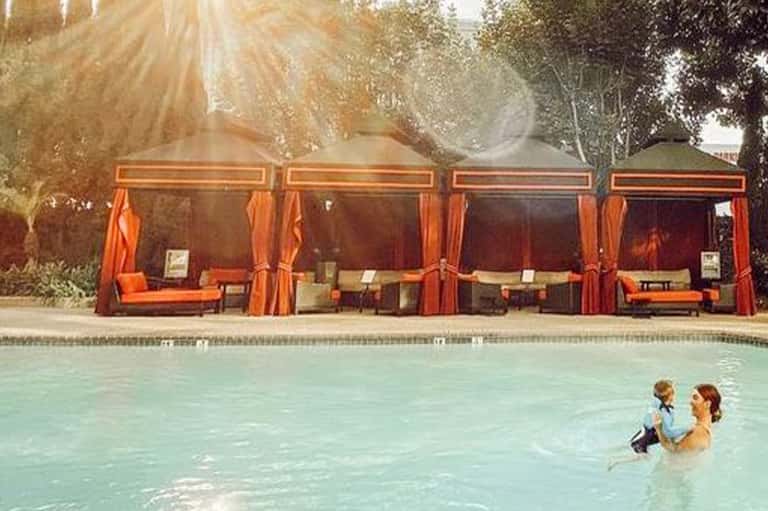 Woman and child wading in resort pool with orange cabanas and palm trees reflected in calm water