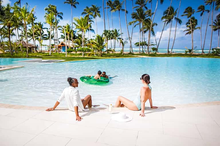 Family relaxing on beach with turquoise lagoon, palm trees, and children on inflatable raft