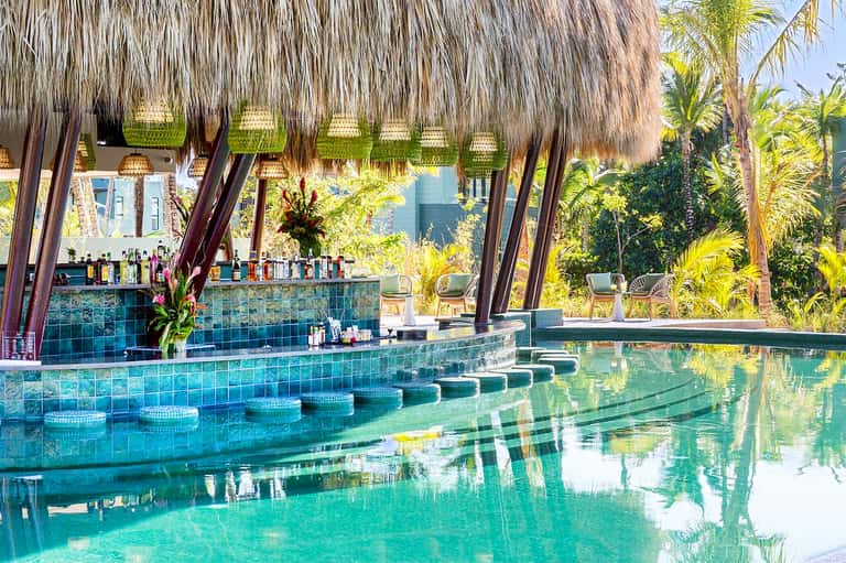 Tropical resort pool bar with thatched roof, turquoise water, palm trees, and bar seating