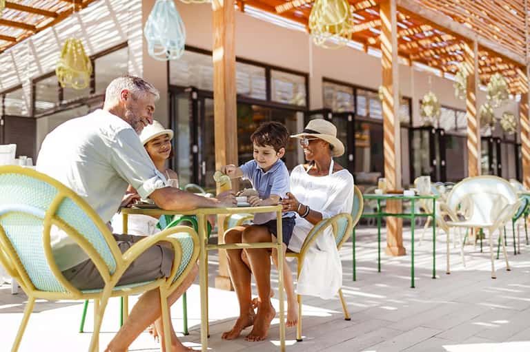 Family dining at resort terrace under wooden pergola with hanging lights and ocean backdrop
