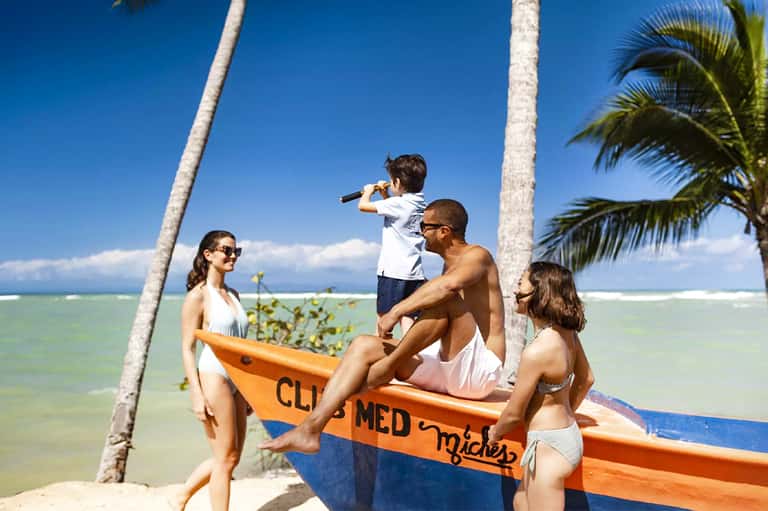 Family with children standing by orange and blue boat on tropical beach with palm trees