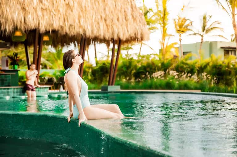 Woman in white swimsuit sitting at edge of tropical infinity pool with thatched-roof structures and palm trees