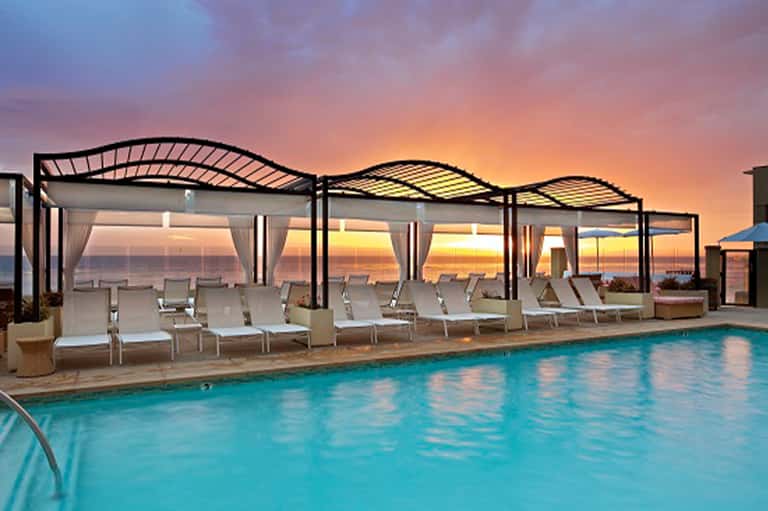 Resort pool at sunset with pergolas, lounge chairs, curtains, and dramatic sky reflection