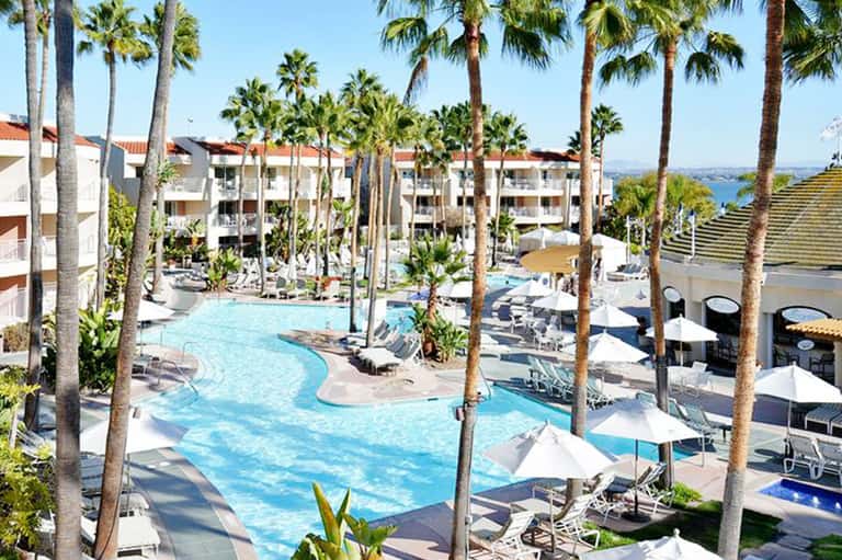 Resort courtyard with curved blue swimming pool, palm trees, and multi-story white buildings under clear sky