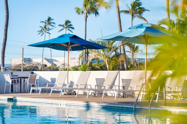 Resort pool deck with blue umbrellas, lounging chairs, and palm trees under clear sky