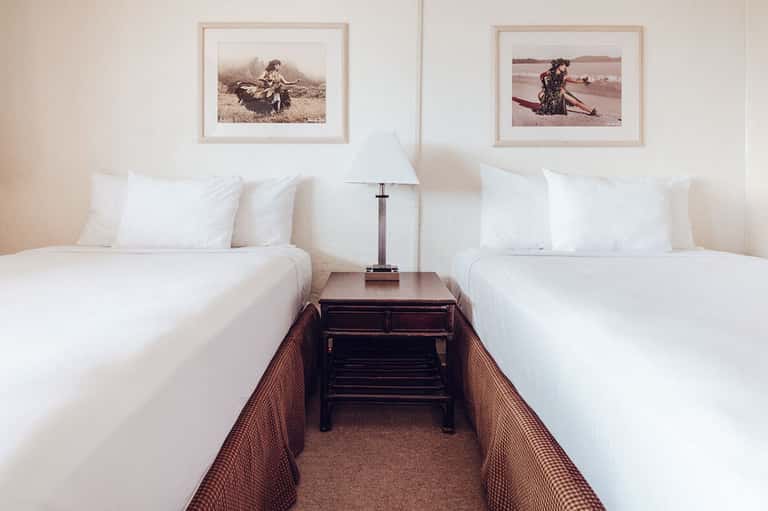Hotel room with two twin beds, white linens, wooden nightstand with lamp, and framed vintage photographs