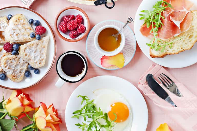 Colorful breakfast spread on pink surface with pastries, eggs, salmon, coffee, and fresh fruit