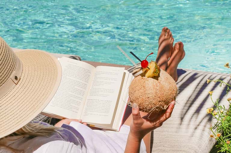 Woman relaxing poolside reading a book while holding a coconut drink