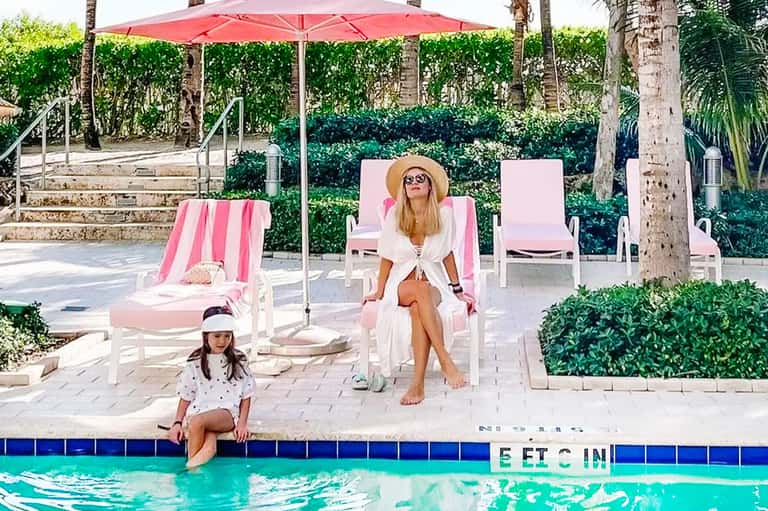 Woman and child relaxing poolside with pink lounge chairs and red umbrella in tropical setting