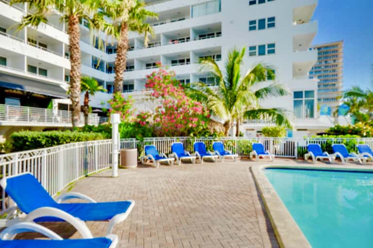 Resort pool deck with blue lounge chairs, palm trees, and multi-story white hotel building